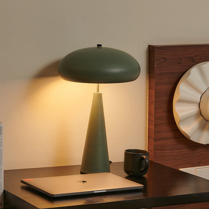 Green mid-century table lamp on a desk with a laptop and mug, wooden wall decor in the background