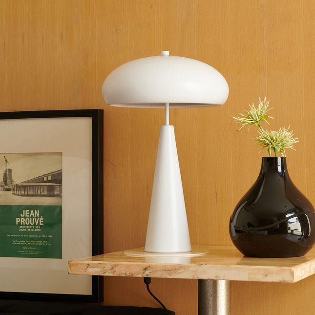 White mid-century table lamp on a wooden surface with a black vase and framed picture in the background.