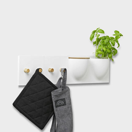 White wall-mounted vertiplants mini plant pot and storage container with hooks holding a black pot holder, gray oven mitt, and a green plant.