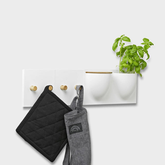 White wall-mounted vertiplants mini plant pot and storage container with hooks holding a black pot holder, gray oven mitt, and a green plant.